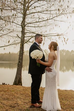 Bride and groom gazing at each other by serene lake holding white rose bouquet under bare tree.