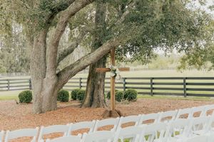 Outdoor wedding ceremony setup with white chairs under a large oak tree with a wooden cross altar.