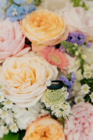 Diamond engagement ring resting on a colorful wedding bouquet with peach roses, pink blooms, and purple flowers.
