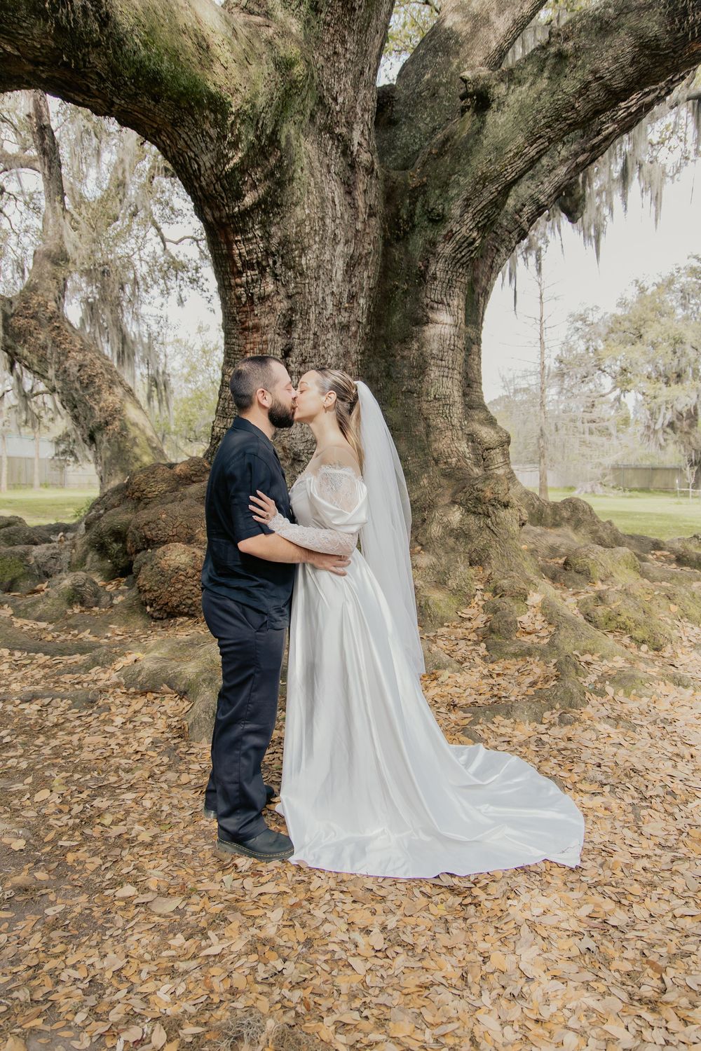 Bride and groom sharing a kiss under a large moss-covered oak tree with fallen autumn leaves.