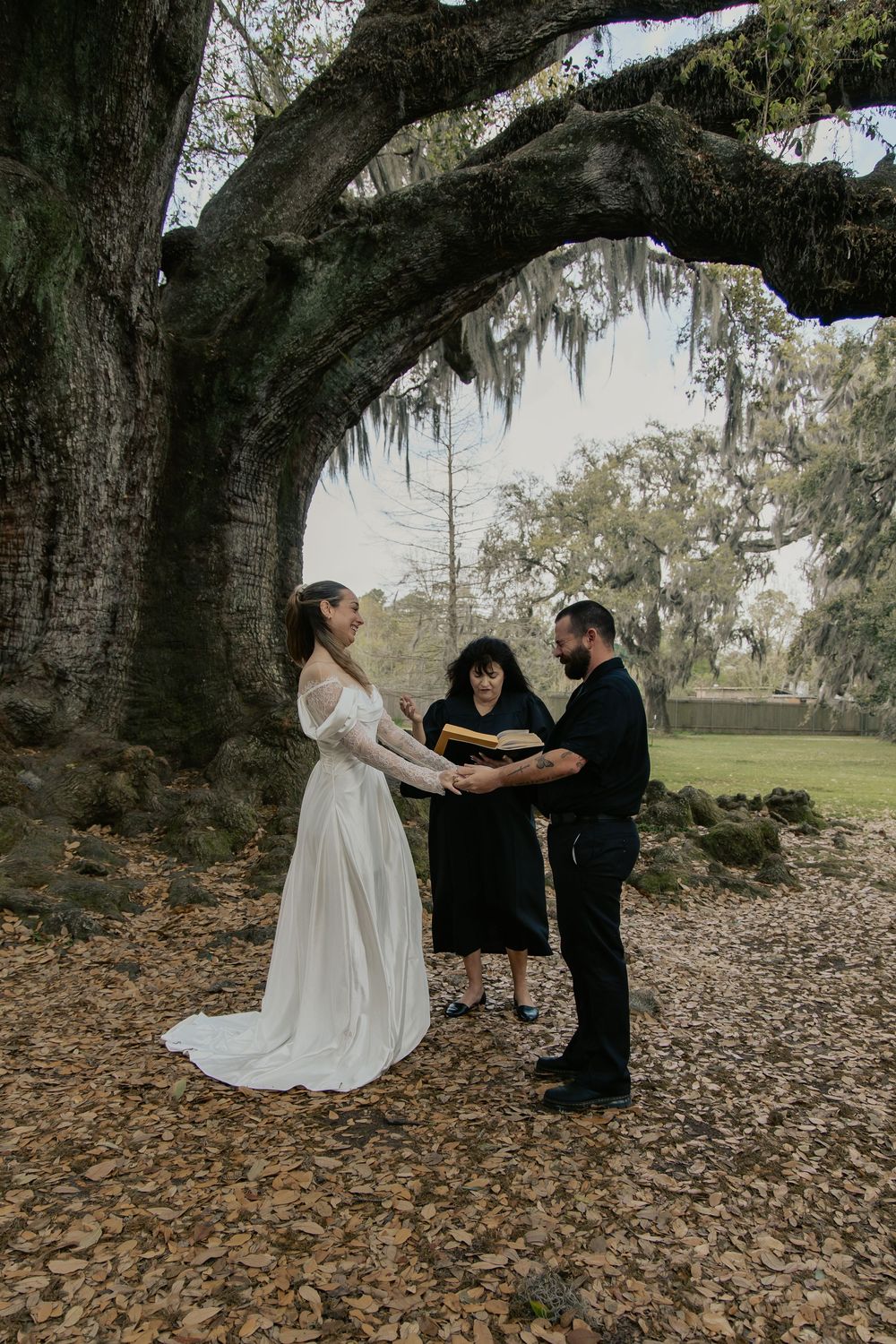 Bride and groom exchanging vows under a large Spanish moss oak tree with a female officiant.