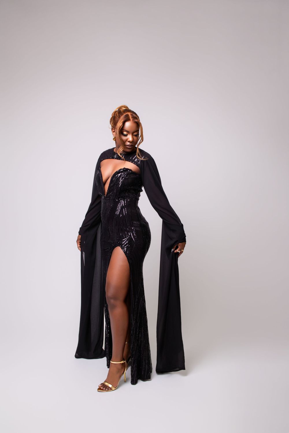 Black woman in elegant black sequin gown with long sleeves and thigh-high slit posing against white background.