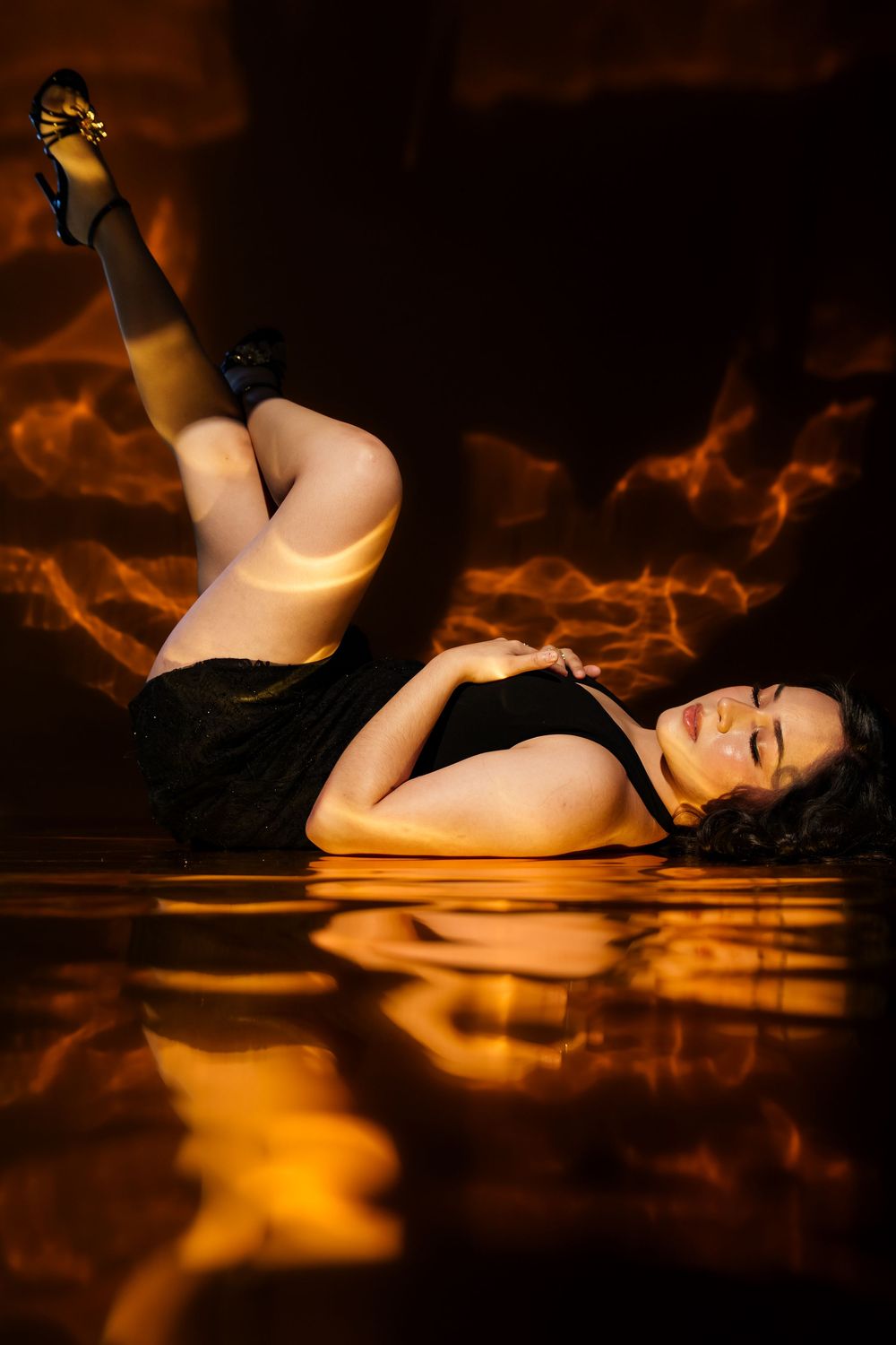 Woman in black dress lying on reflective floor with dramatic fiery orange smoke background and high heels raised.