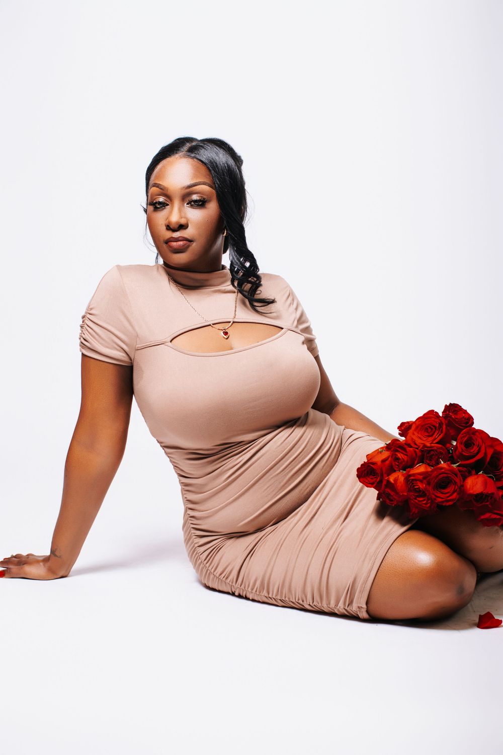A woman in a beige bodycon dress sits gracefully on a white floor holding a bouquet of red roses.