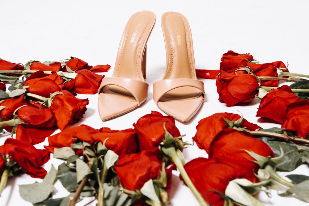 Nude high heel mule sandals surrounded by red roses and petals on a white background.
