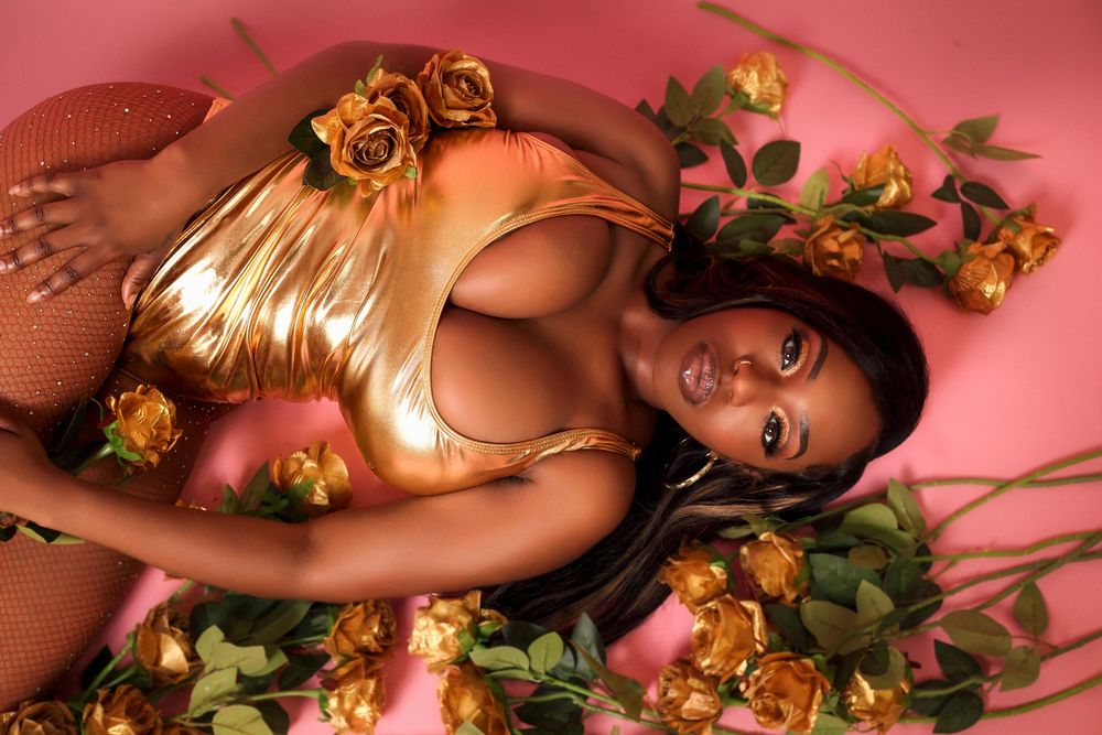 A woman in a gold metallic outfit lies on a pink background surrounded by golden roses and green stems.