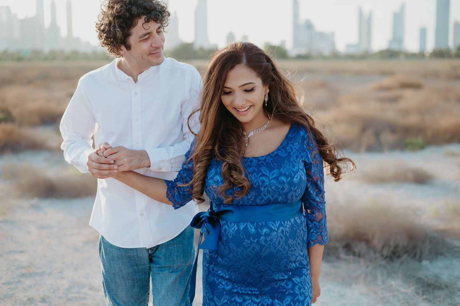 Couple enjoying a maternity photoshoot outdoors, woman in blue lace dress with city skyline in background.