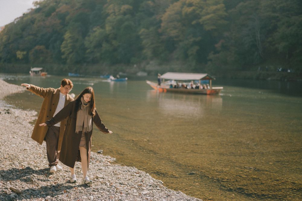 Couple walking along a rocky riverbank in autumn, with traditional boats floating on calm water and colorful foliage in background.