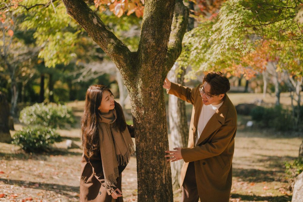 Couple sharing a romantic moment beside a tree in a Japanese autumn park with vibrant red and orange maple leaves.
