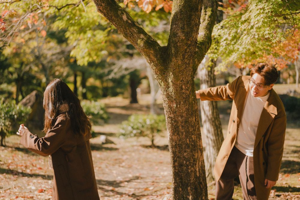 Couple playfully posing around a tree trunk in a sunlit autumn park with golden fallen leaves on the ground.