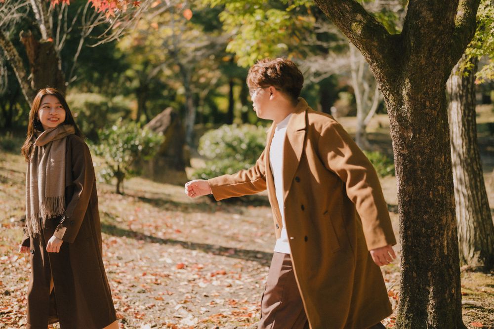 Couple walking through a scenic autumn park with fallen orange leaves and tall trees in warm sunlight.