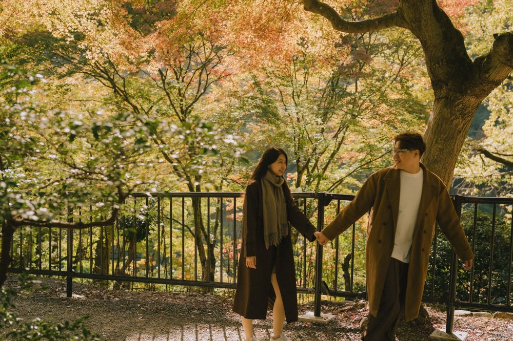 Couple holding hands facing each other beneath stunning autumn maple trees with colorful orange and red leaves.