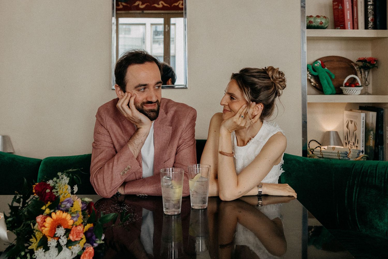 Couple sitting at a dark table with drinks, man in pink blazer and woman in white top, smiling in a cozy room.