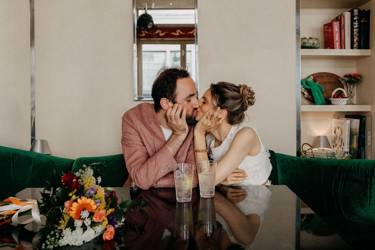 Couple sharing a romantic kiss at a green velvet booth in a stylish bar with colorful flowers and drinks on the table.