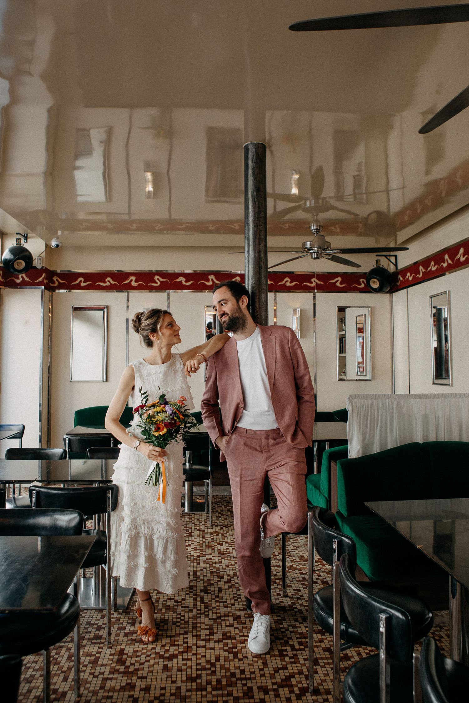 A couple in wedding attire walks through a vintage diner, bride holding colorful bouquet, groom in pink suit.