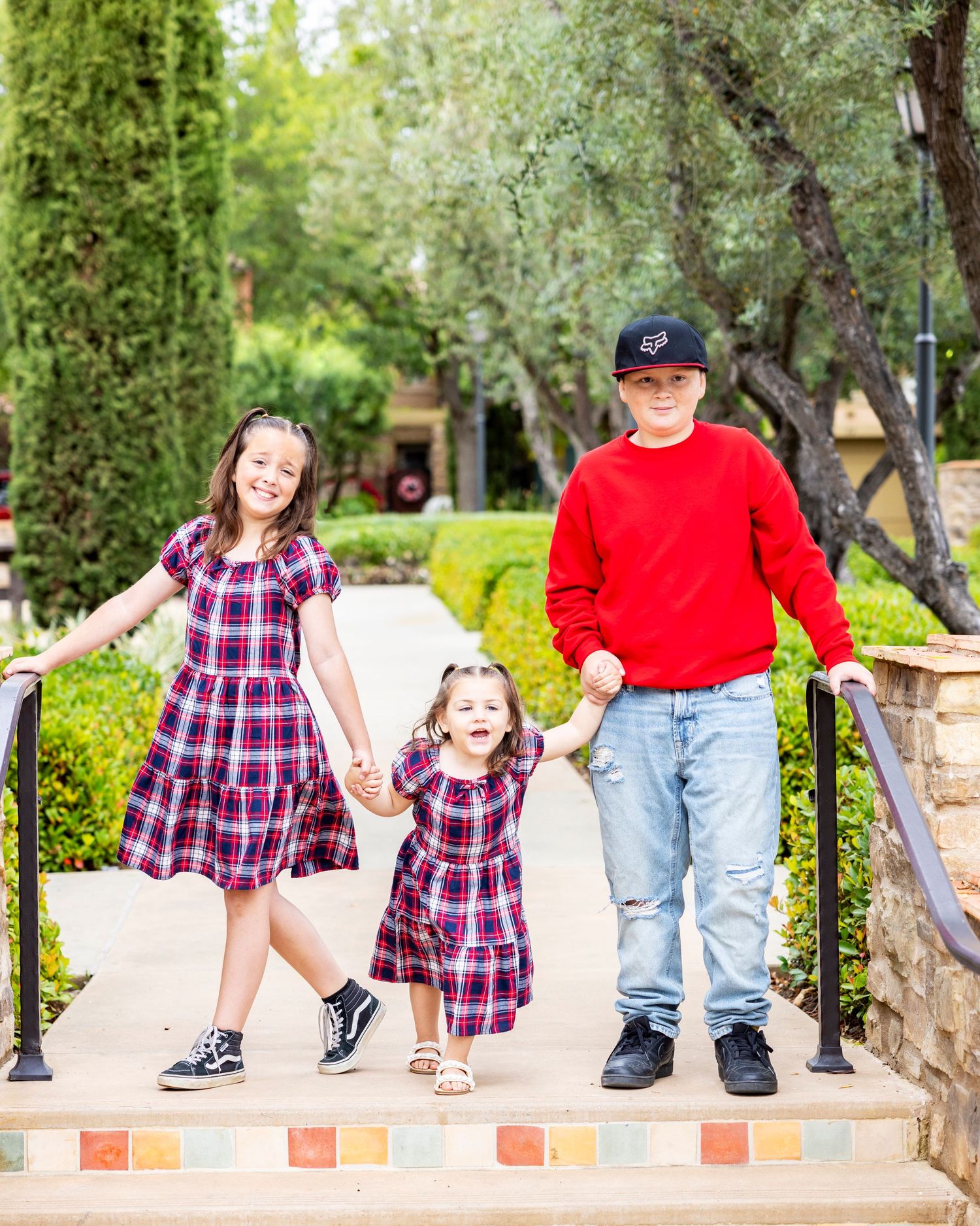 Three children holding hands on decorative steps outdoors, two girls in matching plaid dresses and a boy in red sweater.