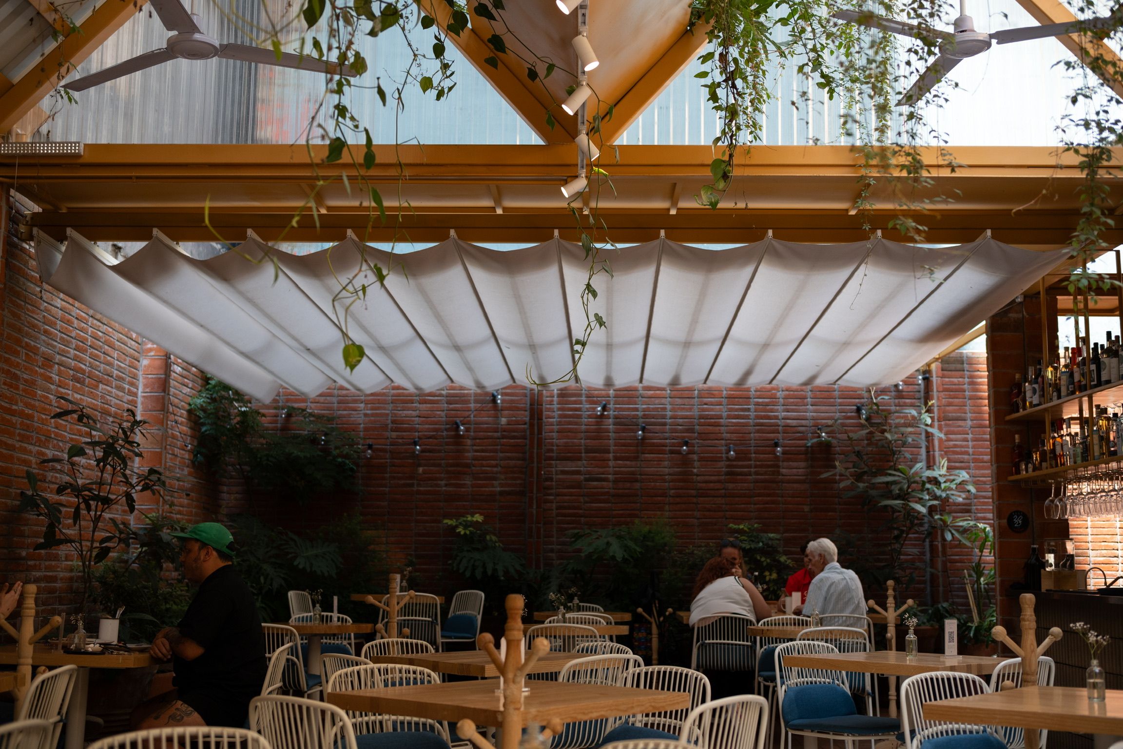 Cozy outdoor patio restaurant with retractable white canopy roof, brick walls, lush plants, and guests dining under warm wooden beams.