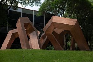 Large abstract Corten steel sculptures with angular geometric forms displayed on green lawn outside modern glass building.