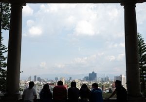Silhouetted visitors admire a panoramic cityscape view from a columned overlook under a partly cloudy sky.