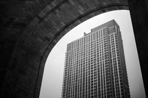 Black and white photo of a tall skyscraper framed by a stone arch, shot from below against an overcast sky.