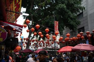 Crowded Chinatown street decorated with red lanterns hanging between trees and buildings during a festive celebration.