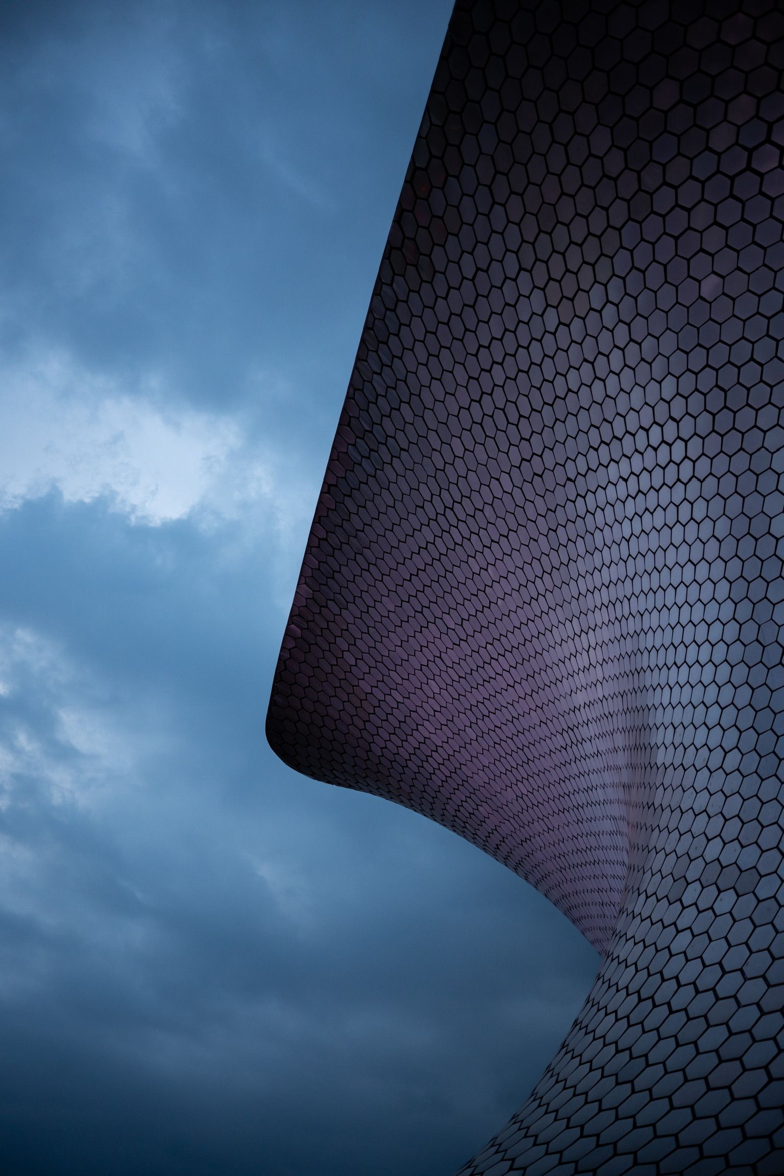 Curved modern building facade with hexagonal pattern against dramatic cloudy blue sky.
