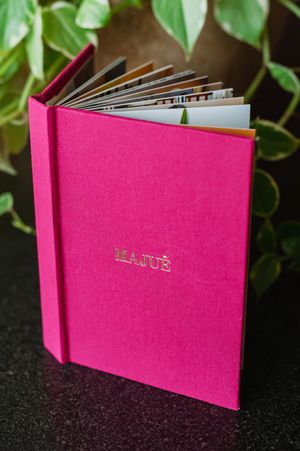 Hot pink hardcover photo album with gold embossed text, filled with photos, displayed against green foliage background.