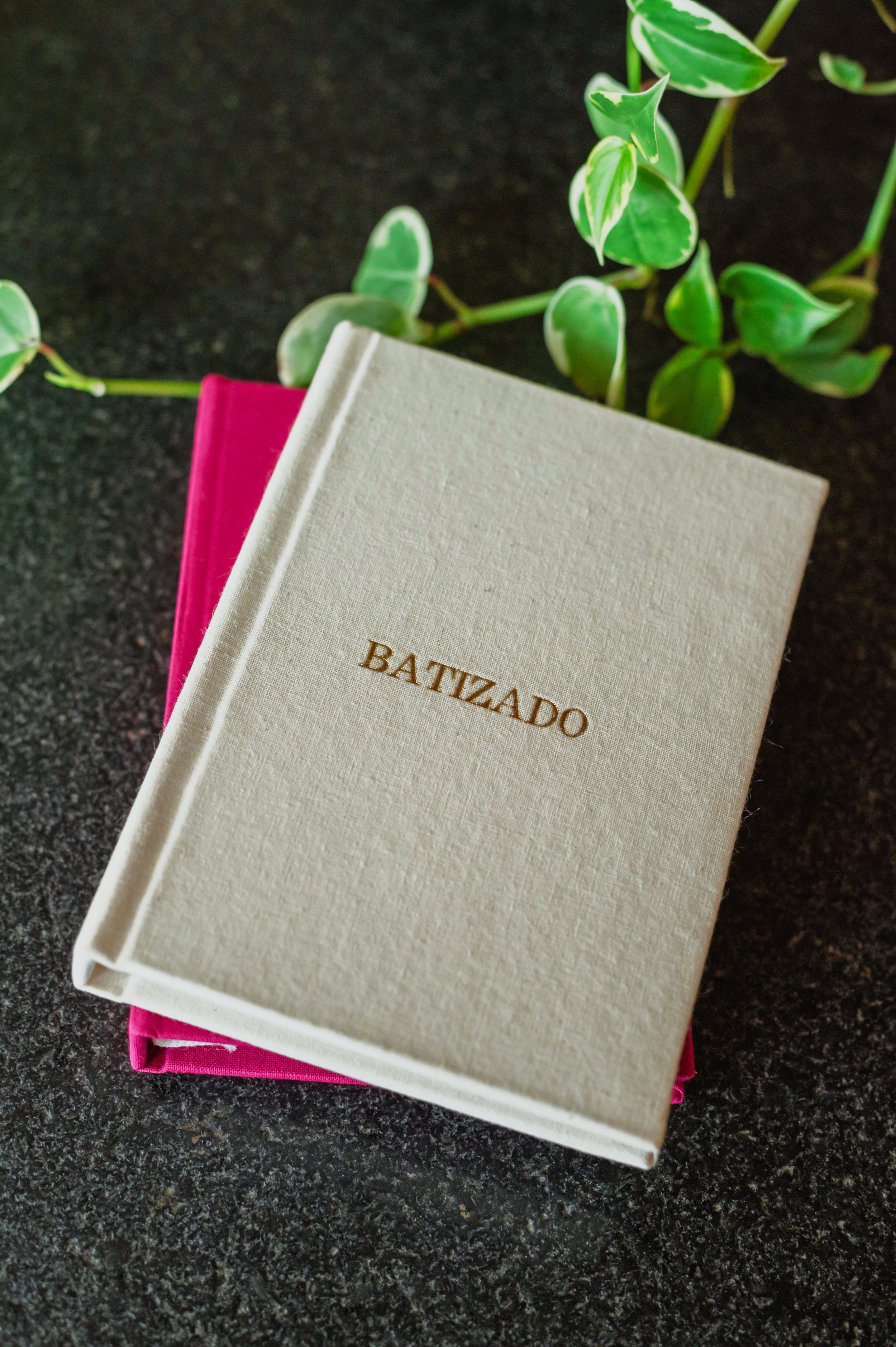 Beige linen hardcover Batizado book resting on a pink book, with green plant leaves on dark surface.