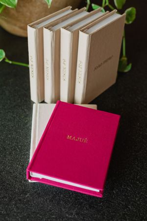 Pink personalized journal labeled MAJUE displayed in front of beige linen hardcover books on dark surface.