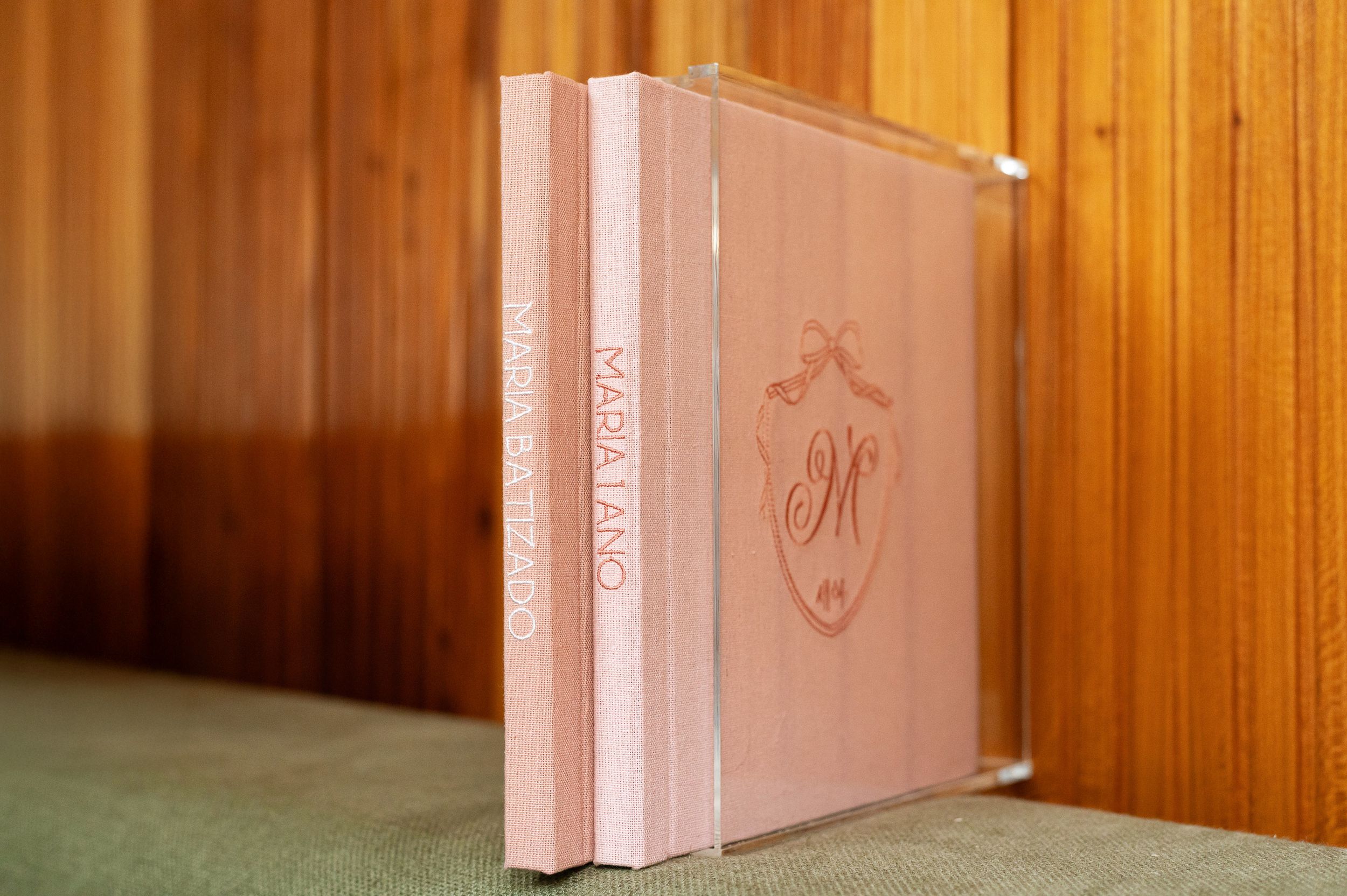 Elegant pink monogrammed wedding album with acrylic case displaying ornate crest design, leaning against wooden wall.
