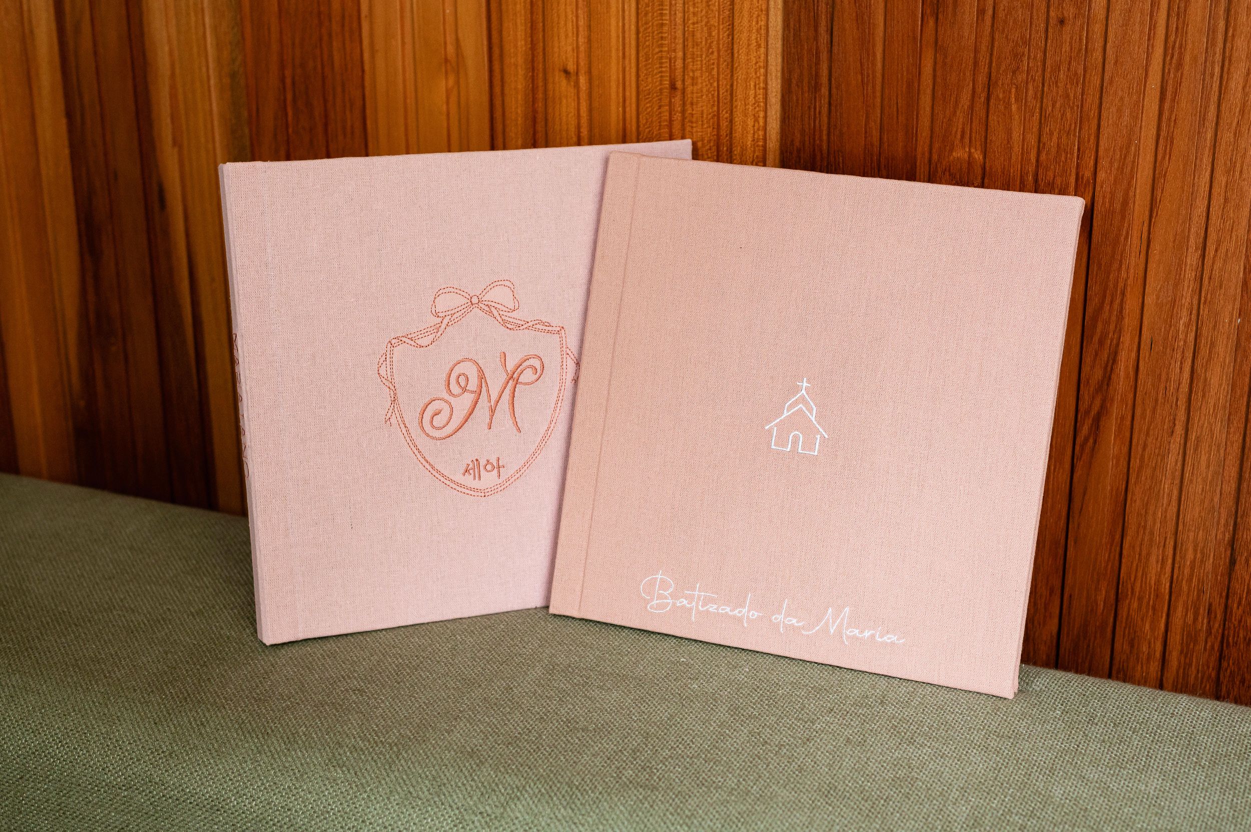 Two pink wedding albums with rose gold foil monogram and church embossed covers displayed against wood paneling.