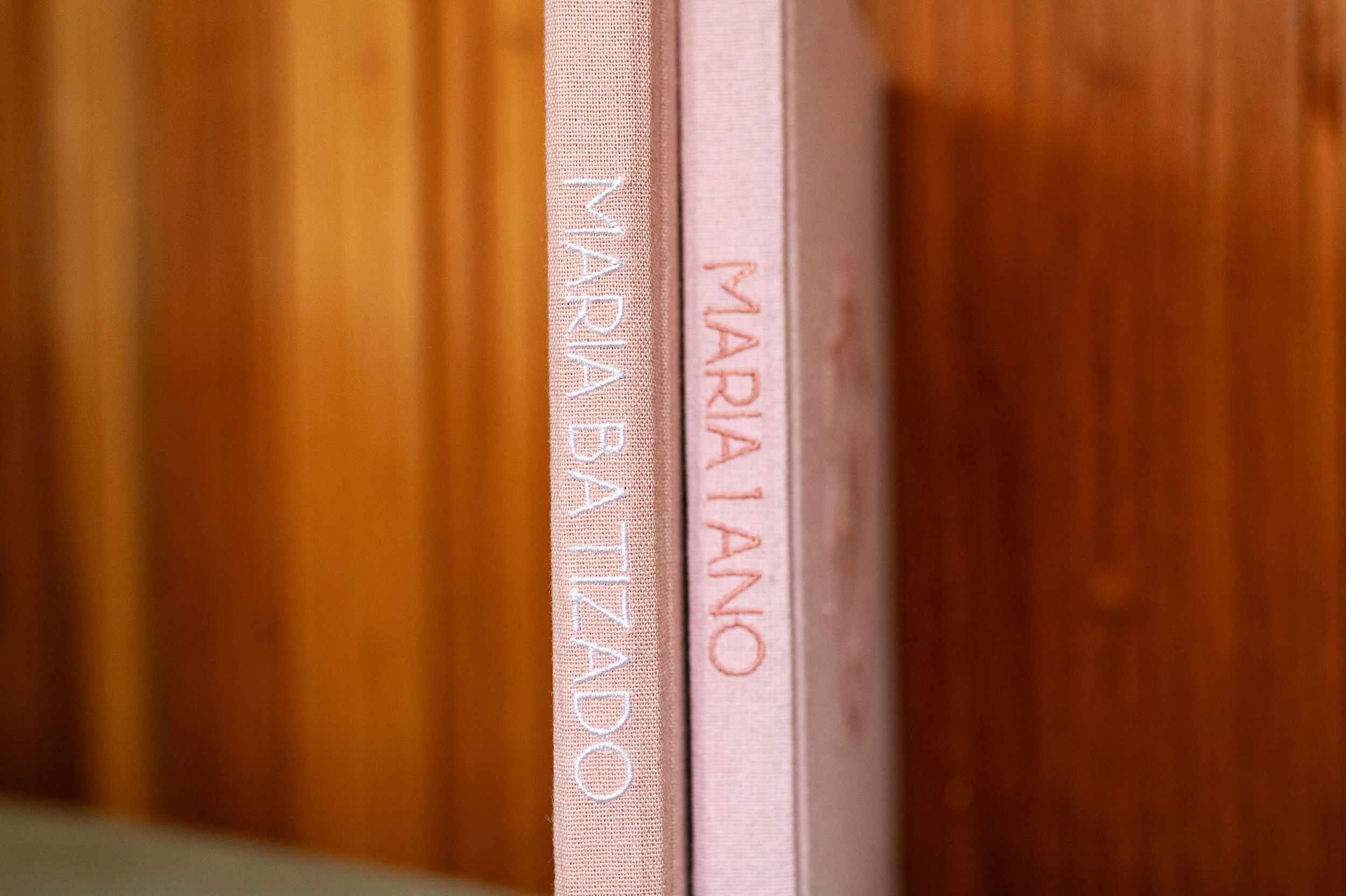 Two pink hardcover books titled Maria Batizado and Maria Lano standing upright on a wooden shelf.
