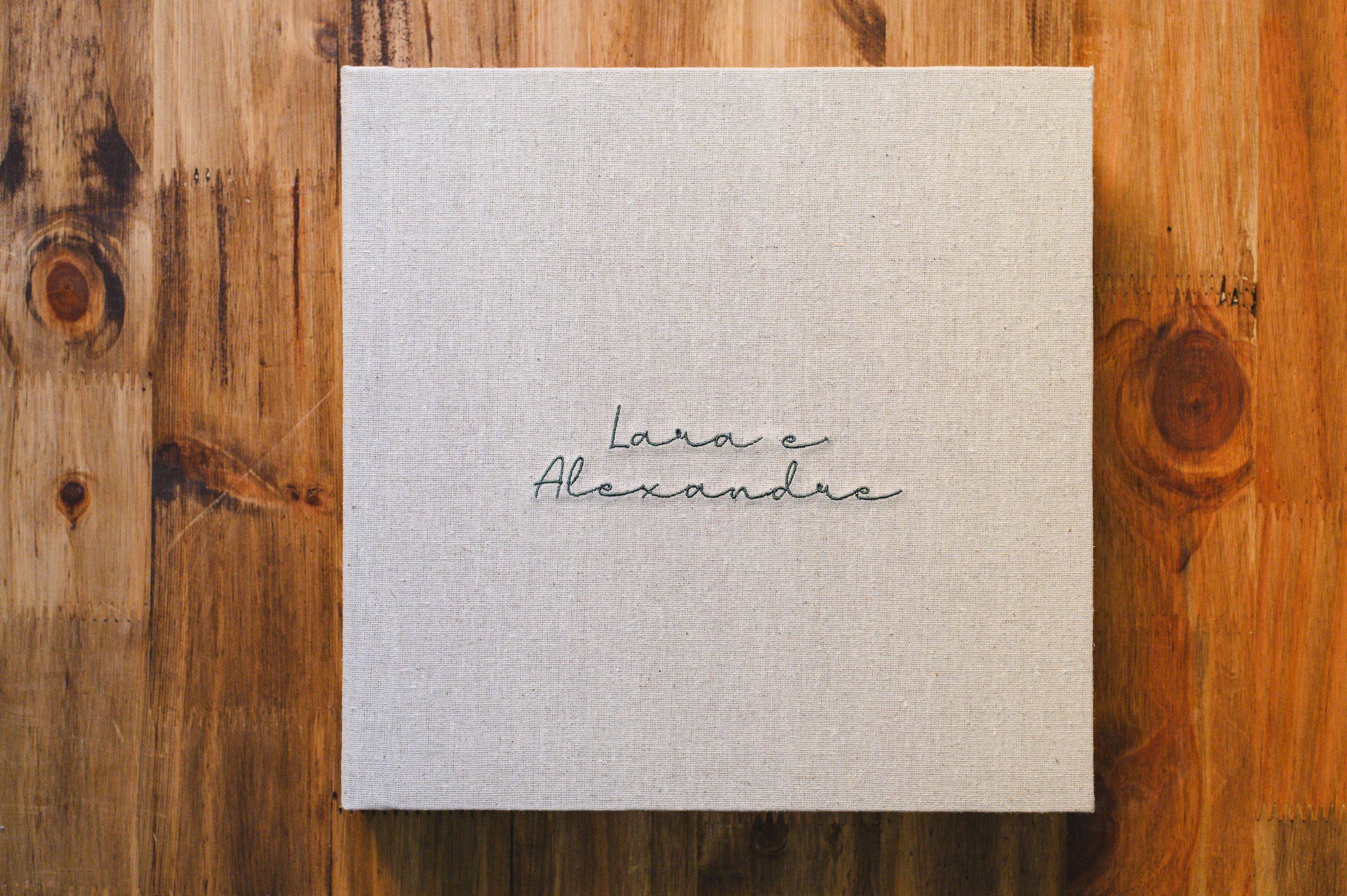 Elegant linen wedding album with cursive names Lara and Alexandre embossed on cover, placed on wooden surface.