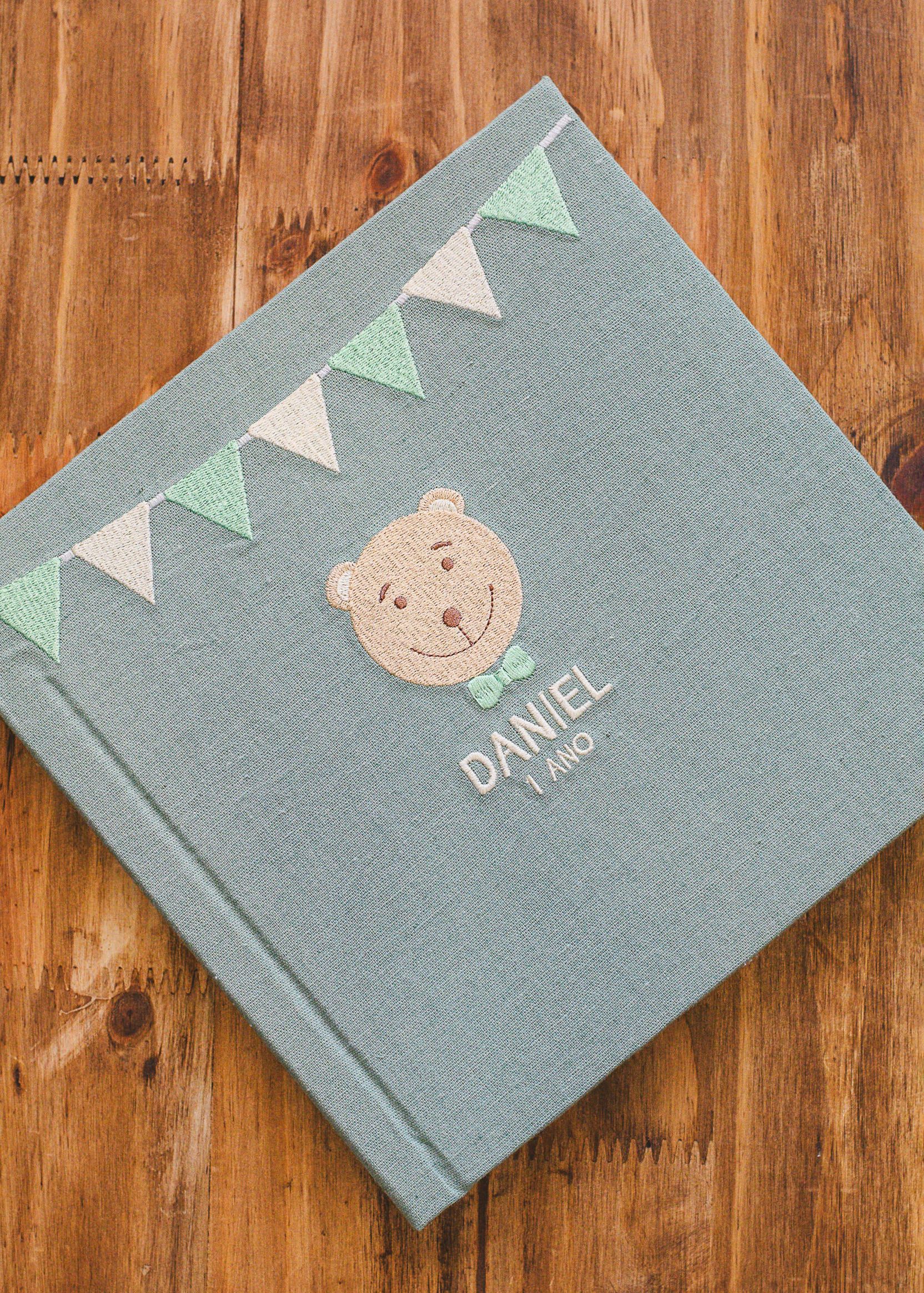 Teal linen baby photo album with bear embroidery, bunting flags, and name Daniel 1 Ano, on wooden surface.