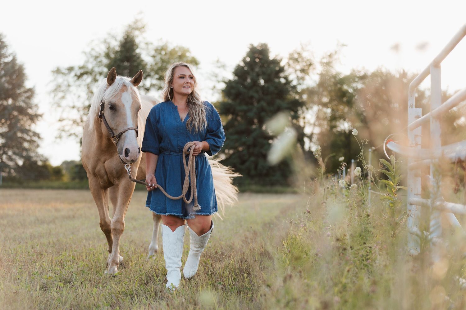 A blonde woman in a blue dress and white boots walks a palomino horse through a sunny green pasture near a fence.
