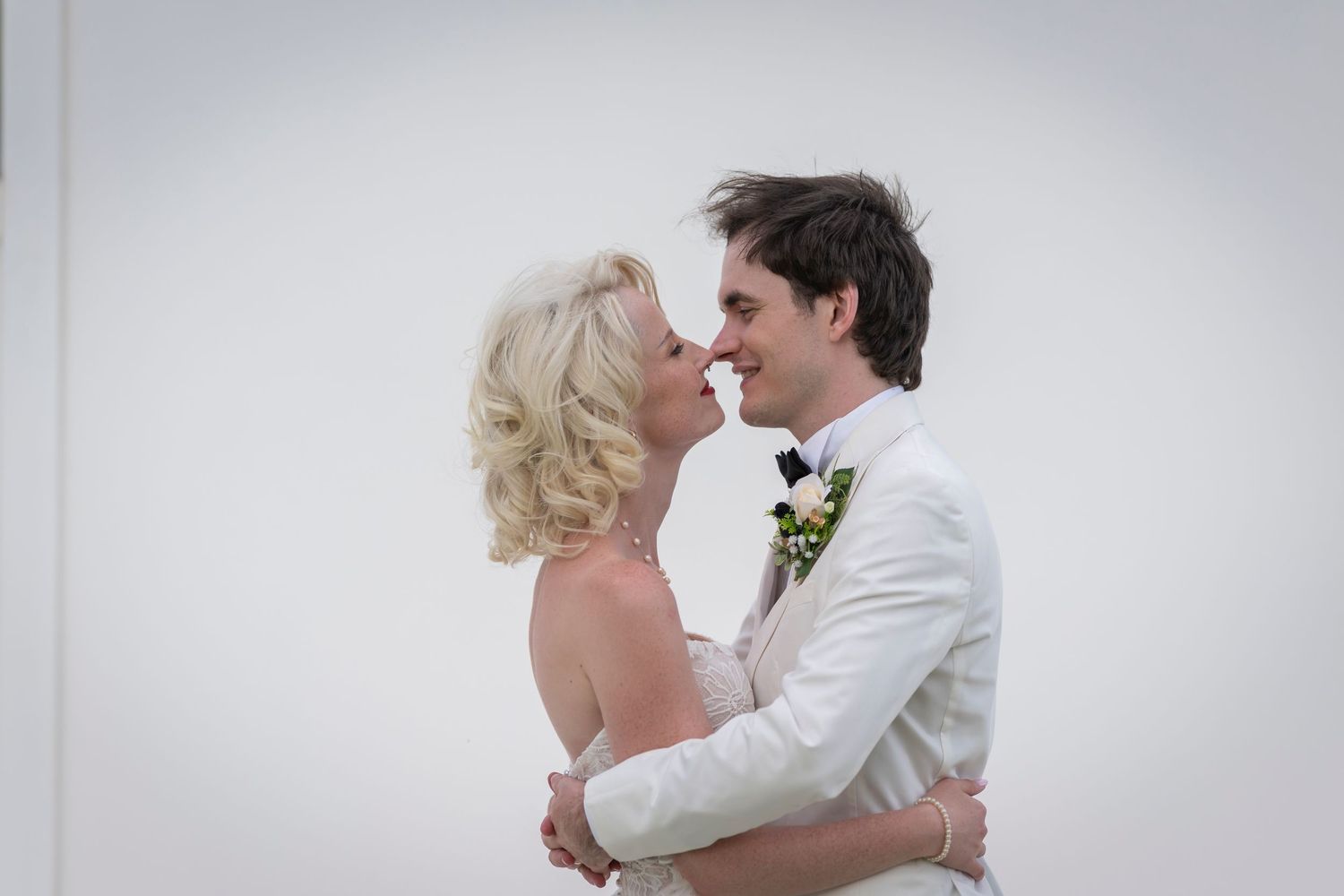 A bride and groom in elegant wedding attire share a romantic embrace, smiling at each other against a white background.