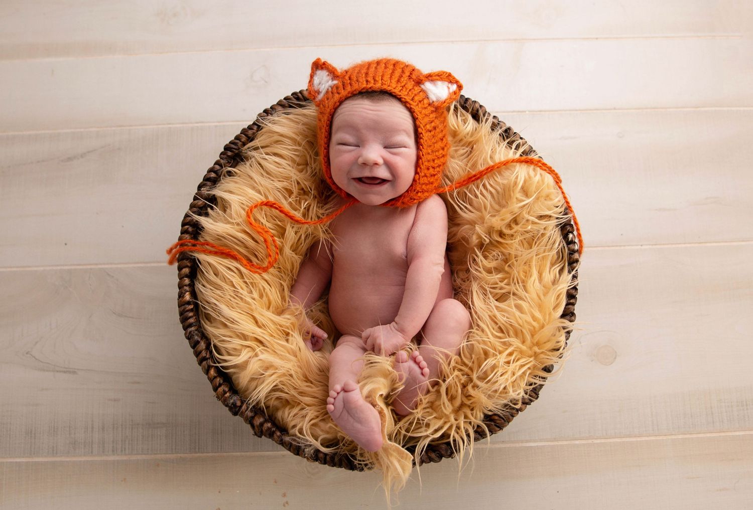 Smiling newborn baby wearing an orange fox knit hat, nestled in a fluffy basket on a light wood floor.