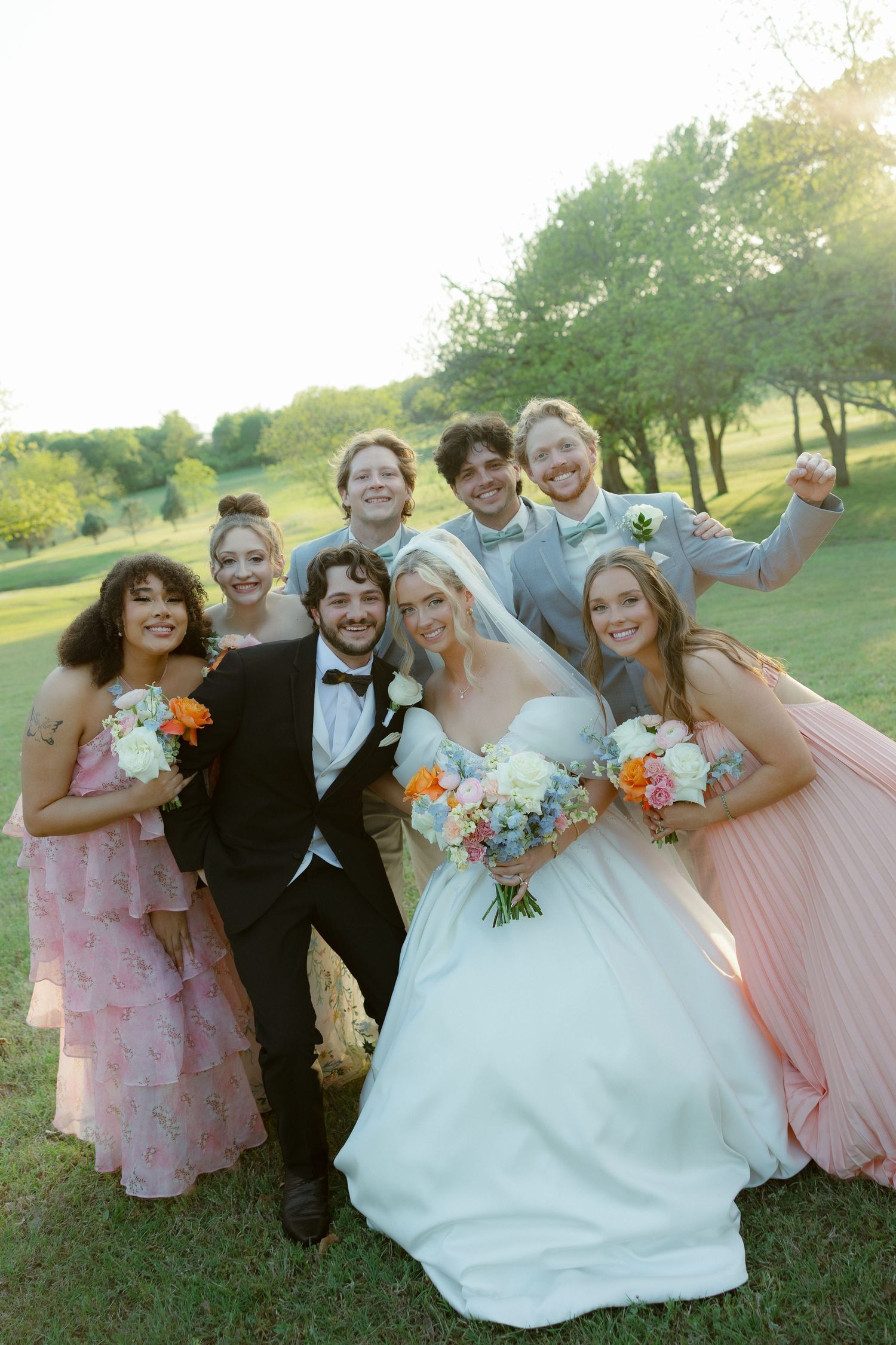 Wedding party poses outdoors on green lawn, bride in white gown holding colorful bouquet with bridesmaids in pink and groomsmen in gray.