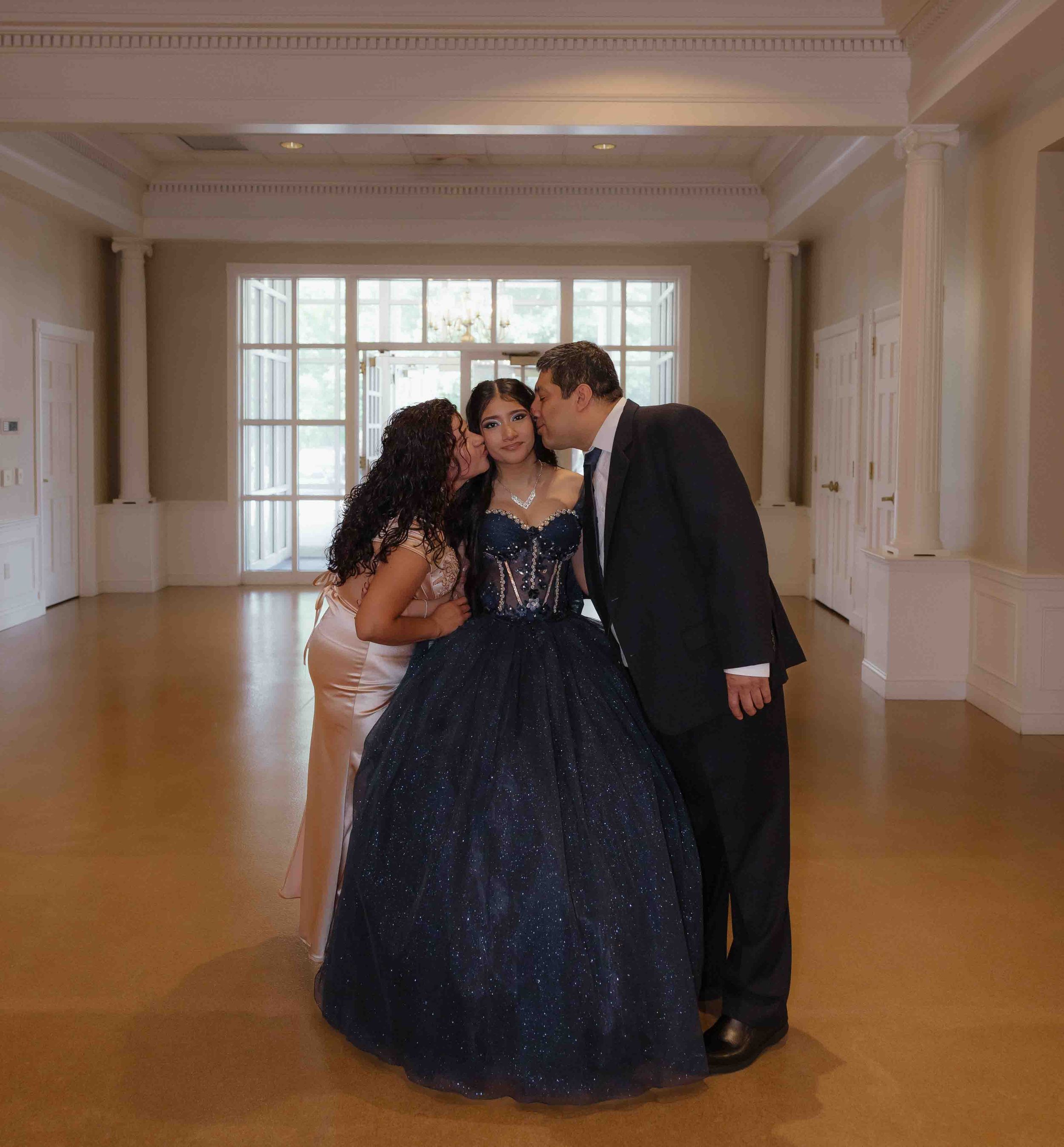 Young woman in navy blue quinceañera ball gown being kissed by her parents in an elegant ballroom.