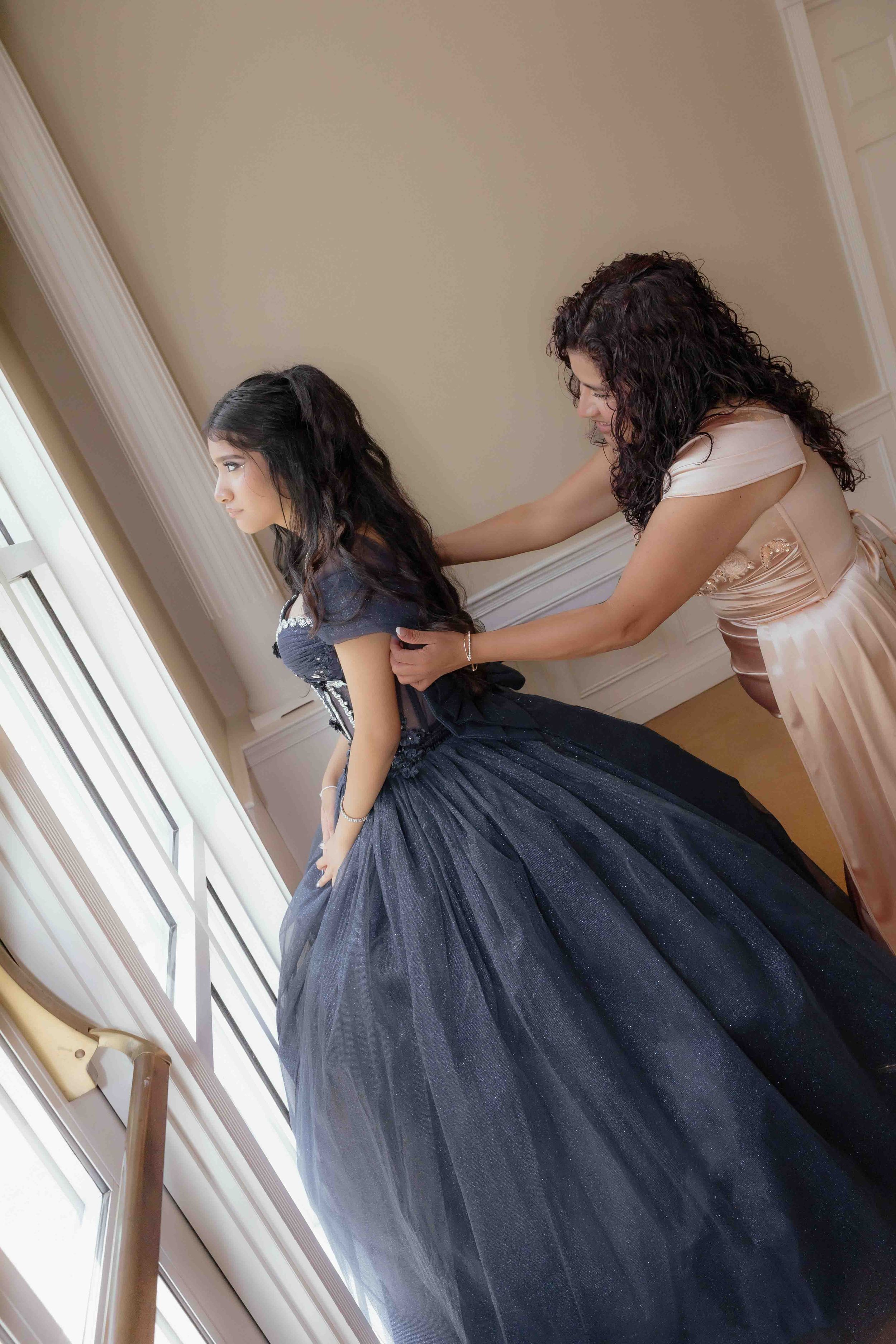 Woman helping a young girl in a navy blue quinceañera gown with her dress near a bright window.