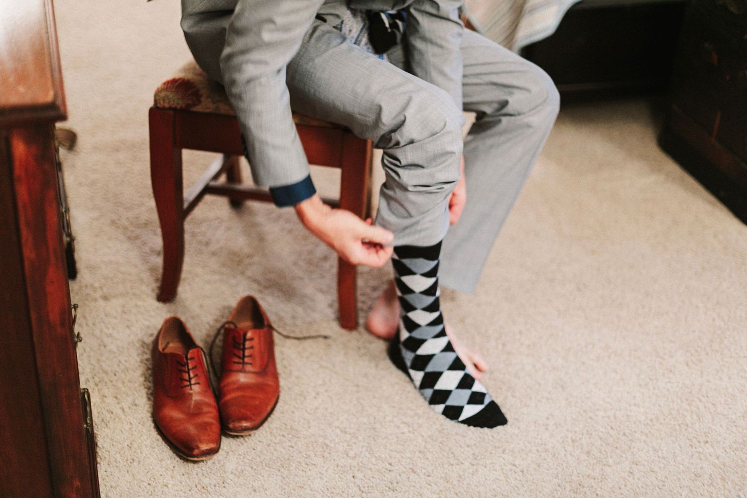 Groom in gray suit putting on black and white argyle socks beside brown leather dress shoes on carpet.