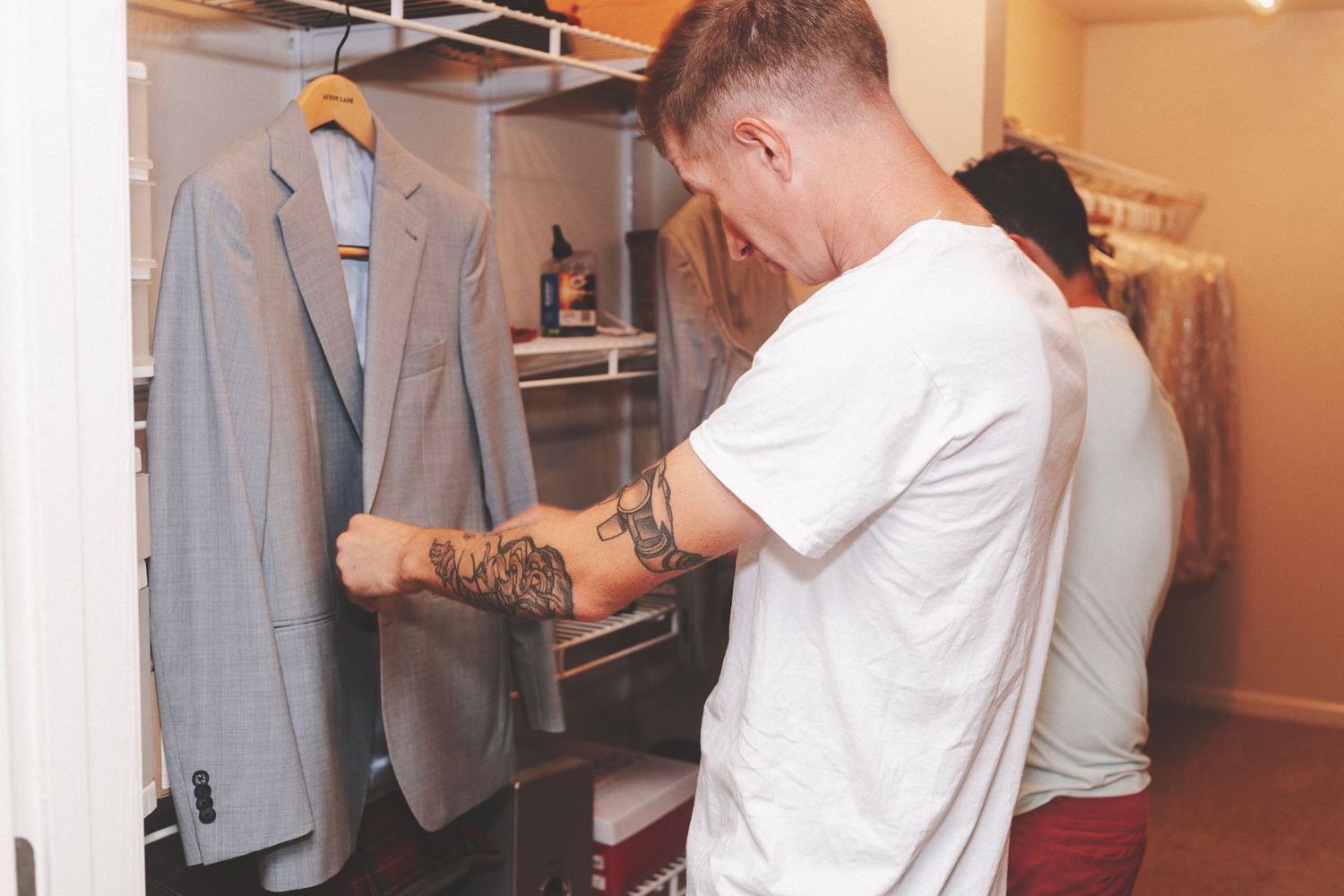 Tattooed man browsing suits in a closet, selecting formal wear with a gray blazer hanging nearby.