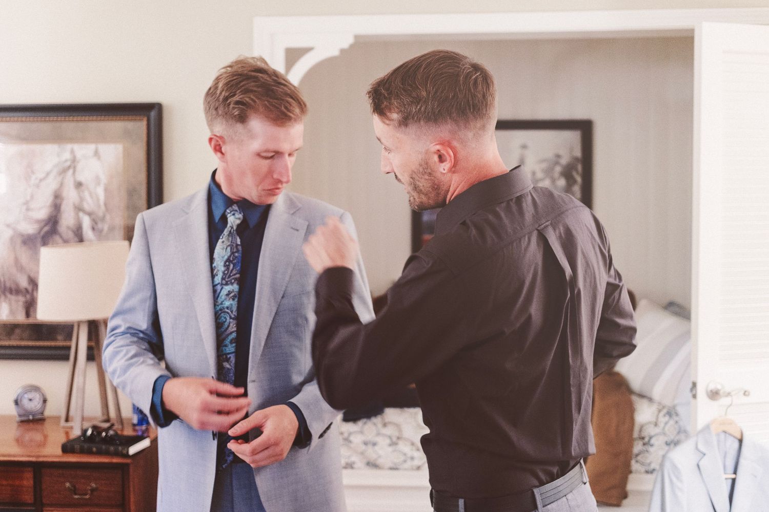 Two men in formal attire, one in a gray suit with tie, getting dressed with assistance indoors.