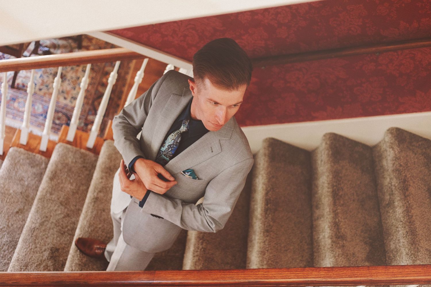 A young man in a gray suit adjusts his cufflinks while descending carpeted stairs indoors.