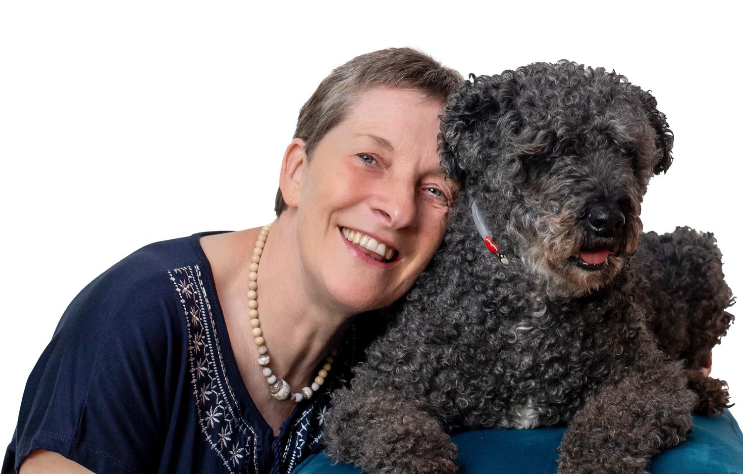 Smiling woman with pearl necklace posing cheek-to-cheek with a fluffy black poodle against white background.