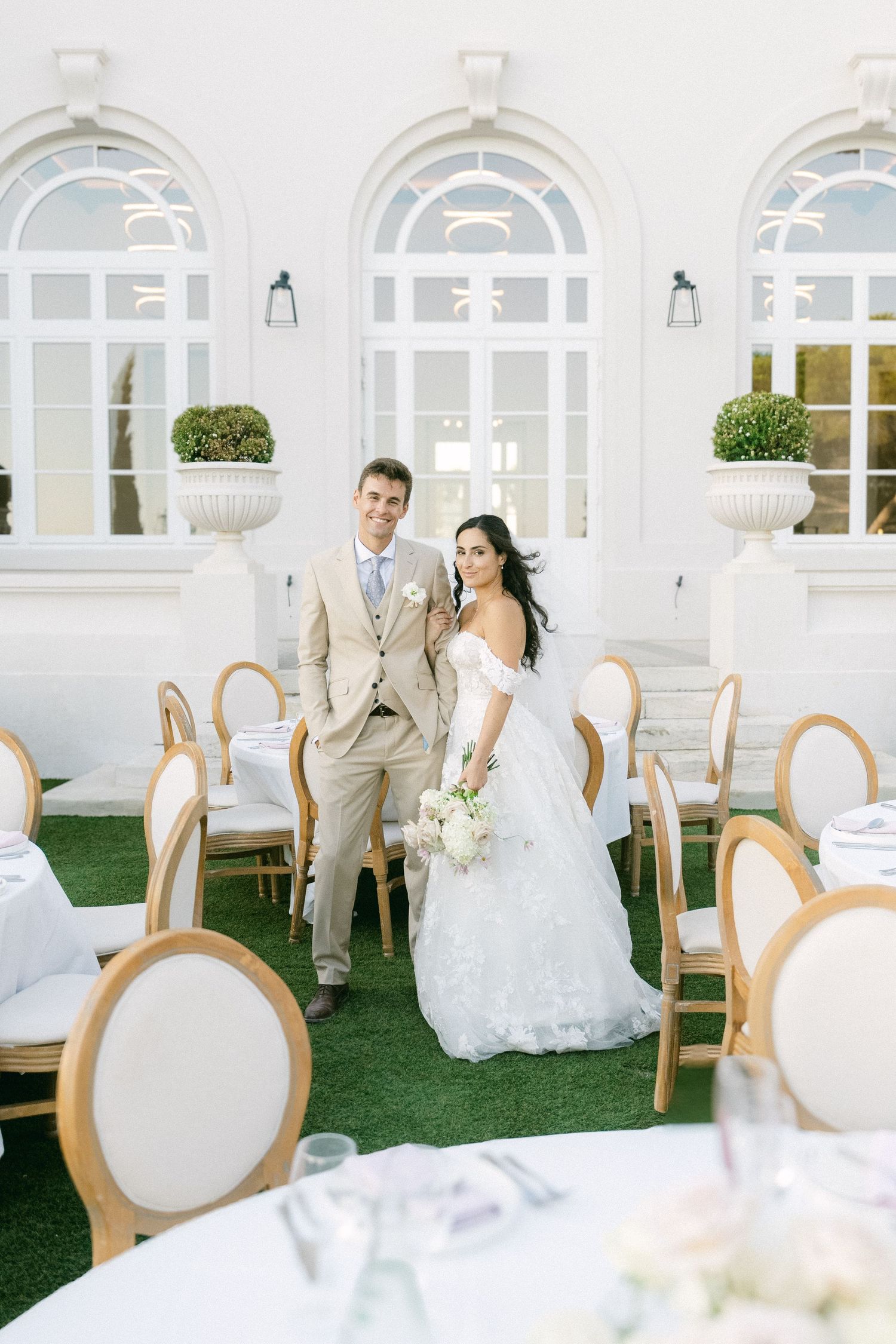 Bride and groom posing together at elegant outdoor wedding venue with white chairs and lush greenery.