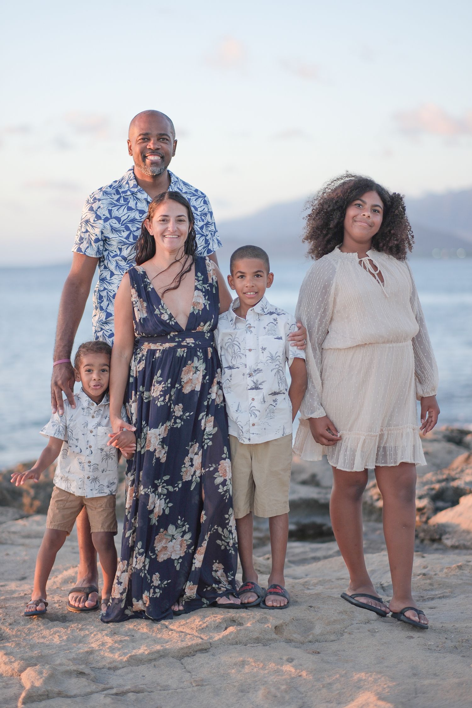 Family of five posing together on rocky beach at sunset, with ocean and mountains in background.