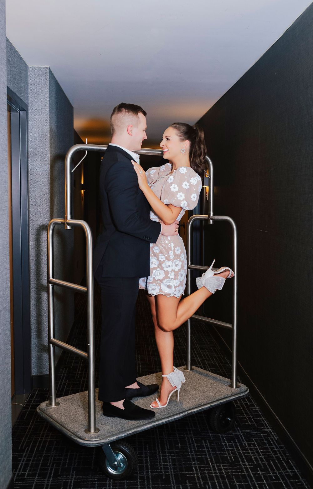 Couple posing romantically on a hotel luggage cart in a hallway, wearing formal attire.