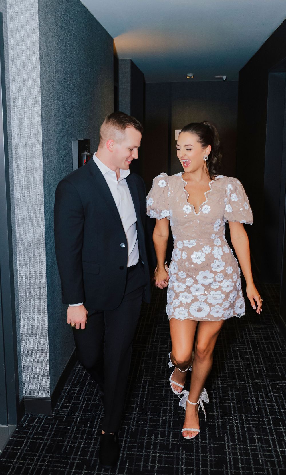 Couple walking hand in hand down a hallway, man in navy suit, woman in white floral mini dress with heels.