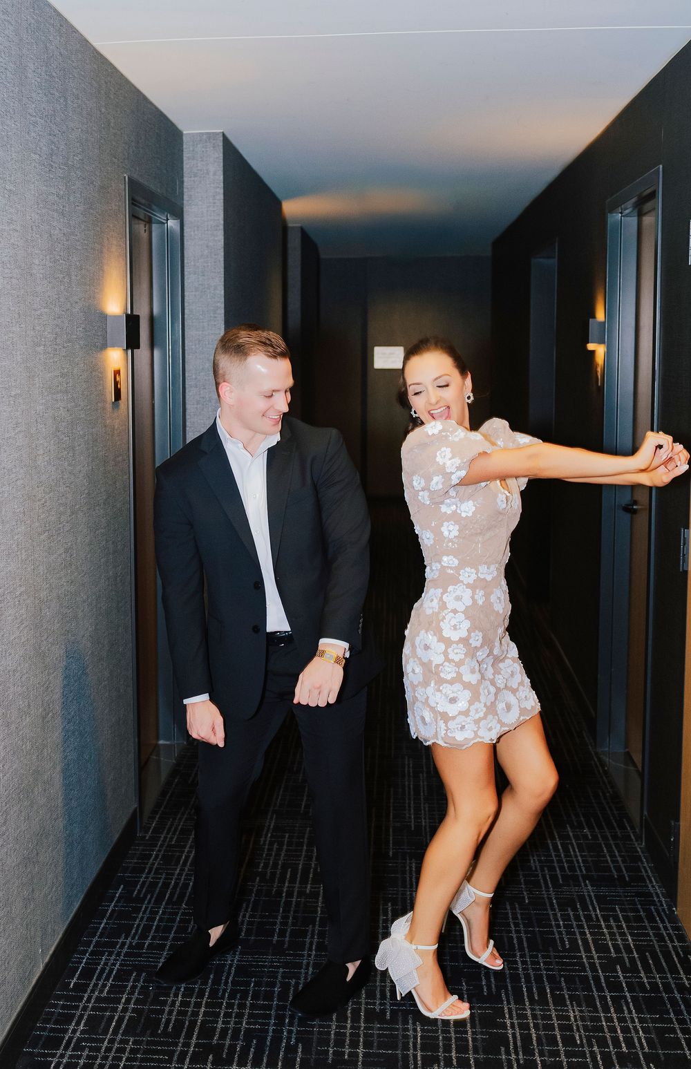 A couple dances joyfully in a modern hotel hallway, man in black suit, woman in floral mini dress with heels.